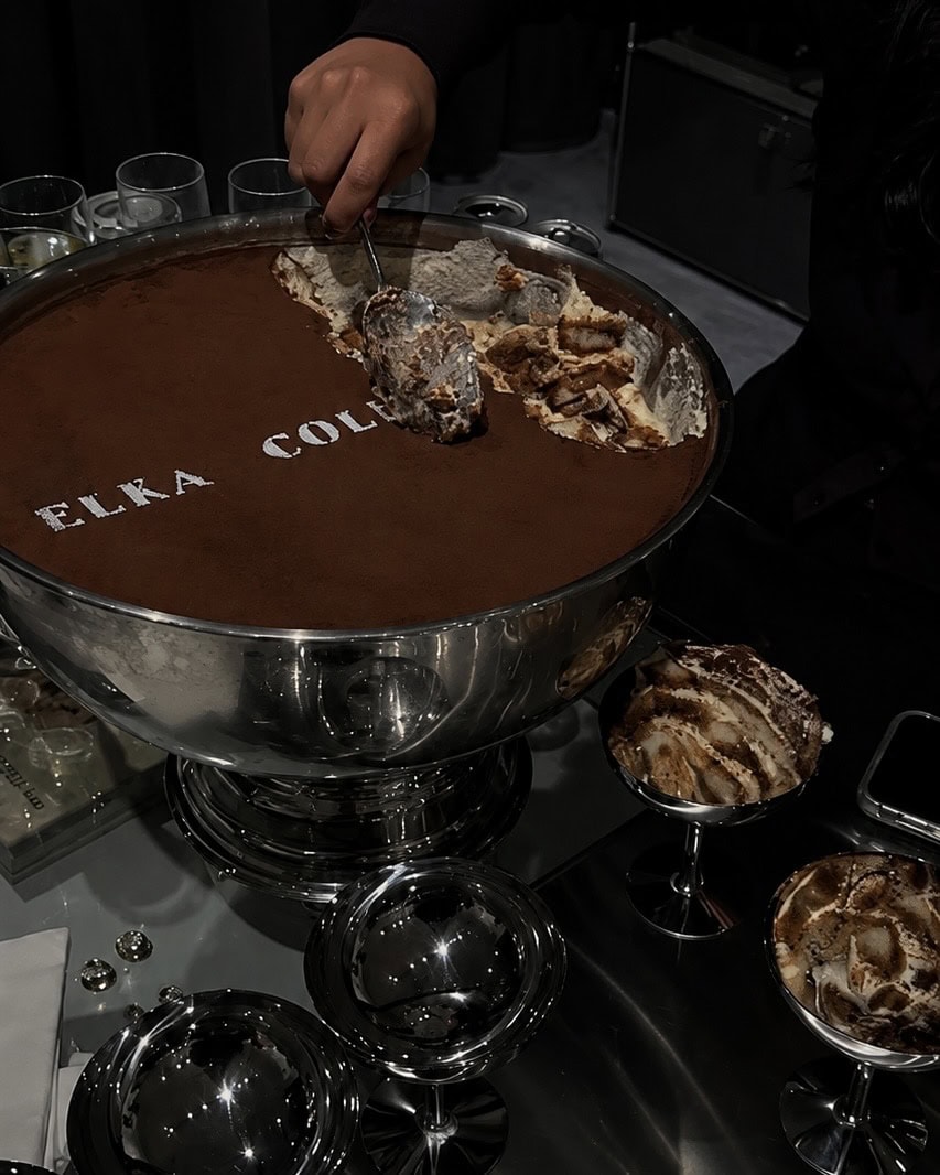 A large silver punch bowl of tiramisu dusted with cocoa powder being served at an event, with individual silver coupe glasses of tiramisu arranged around it on a dark reflective surface.