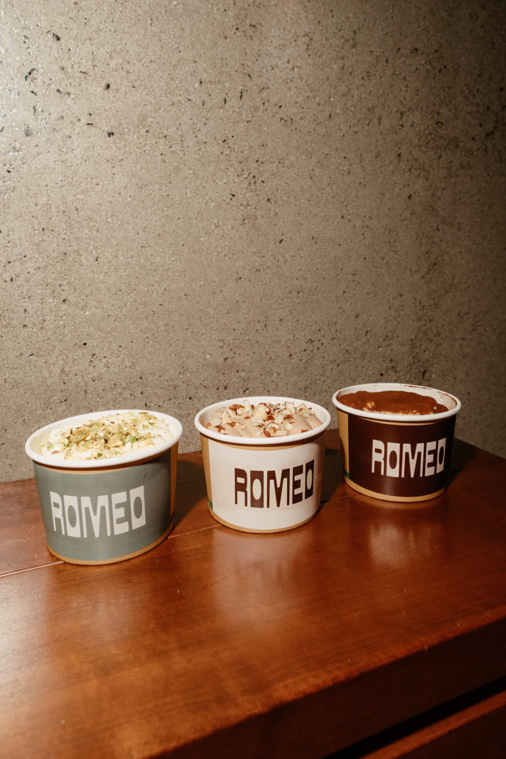 Three Romeo-branded tiramisu cups lined up on a timber surface in Adelaide, each topped with different garnishes — crushed pistachios, toasted almonds, and cocoa powder — against a concrete wall backdrop.