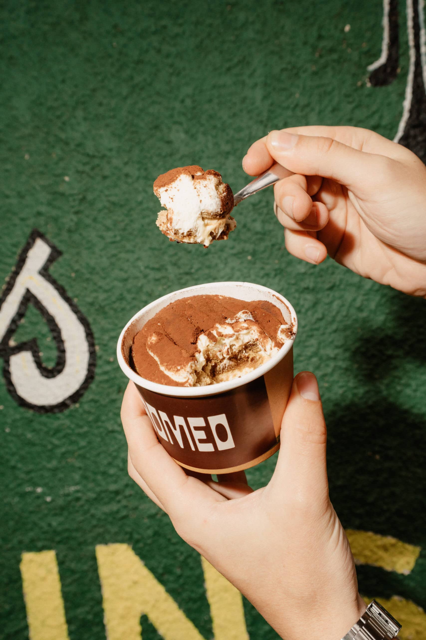Person scoops tiramisu from a Romeo-branded cup dusted with cocoa powder, against a green textured wall with street art details in Adelaide.