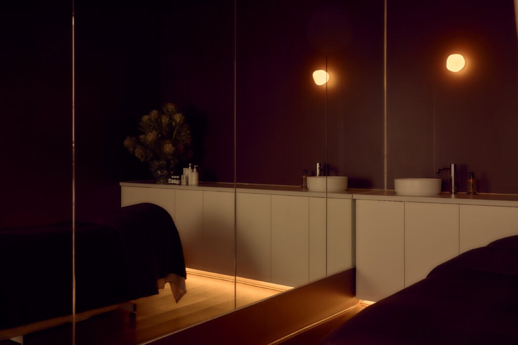 Moody facial treatment room at Fluffy Skin in Cremorne, featuring burgundy walls, warm globe lighting, mirrored panels, white basins and a treatment bed draped in dark towels.