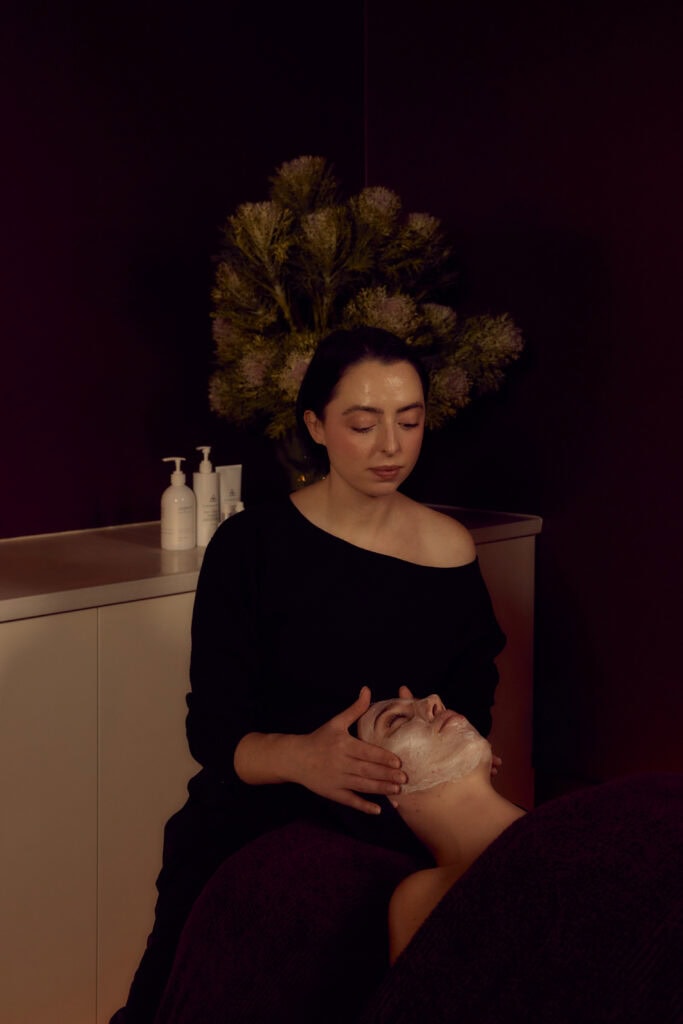 Close-up of a Fluffy Skin facial in Cremorne, showing a therapist applying cleanser to a client's face against a backdrop of burgundy walls and warm lighting.