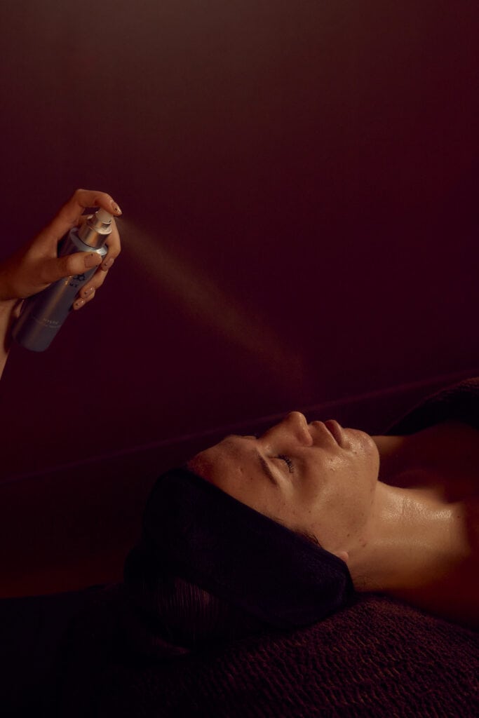 A client lies with eyes closed during a facial at Fluffy Skin in Cremorne, as a therapist sprays a fine mist over her face against a deep burgundy backdrop.