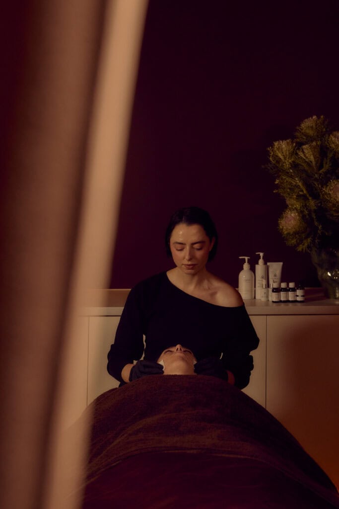 Skin therapist tending to a client during a facial at Fluffy Skin in Cremorne, with dark burgundy towels, botanical arrangements and skincare products in the background.