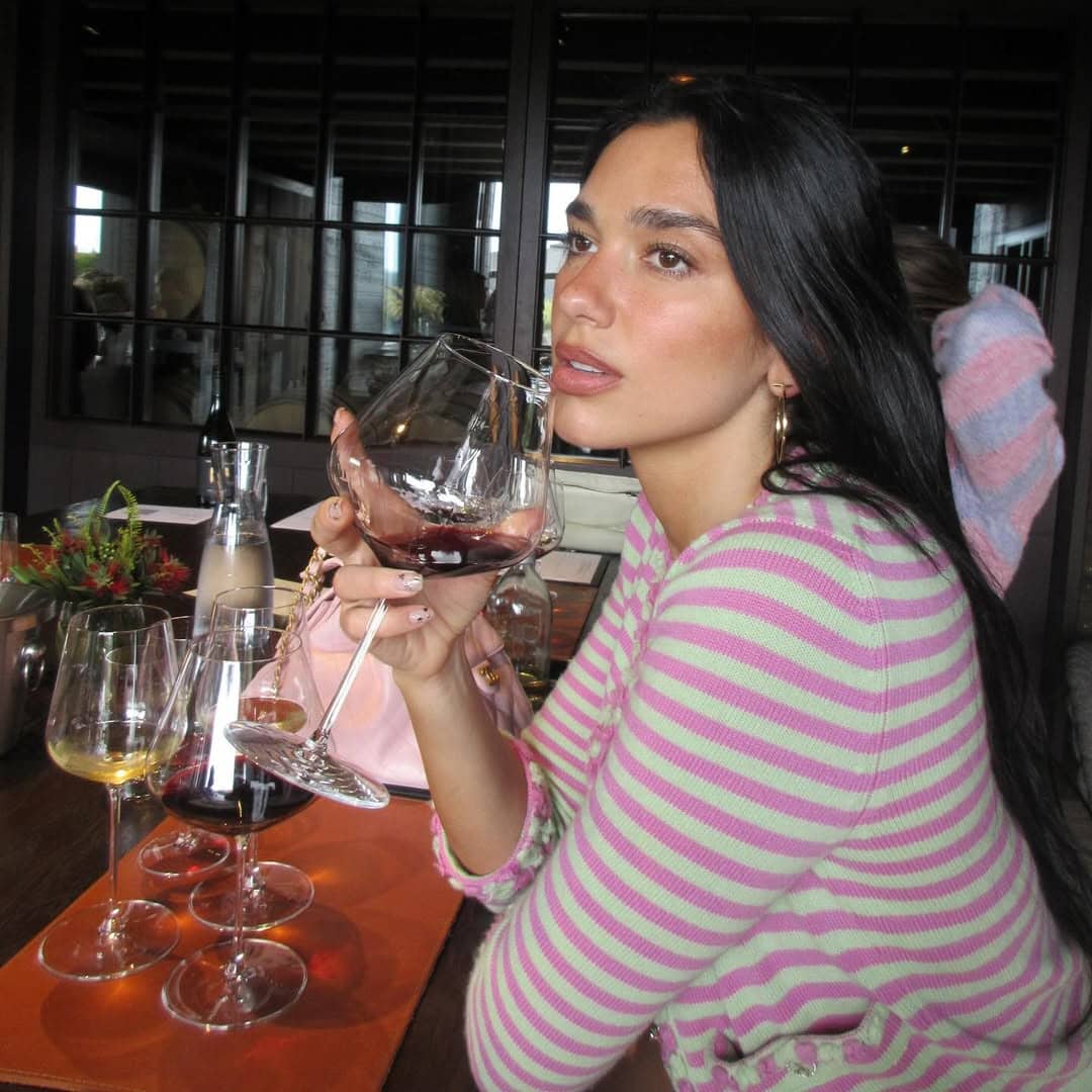 dua lipa in a striped knit top holding a glass of red wine and looking into the distance