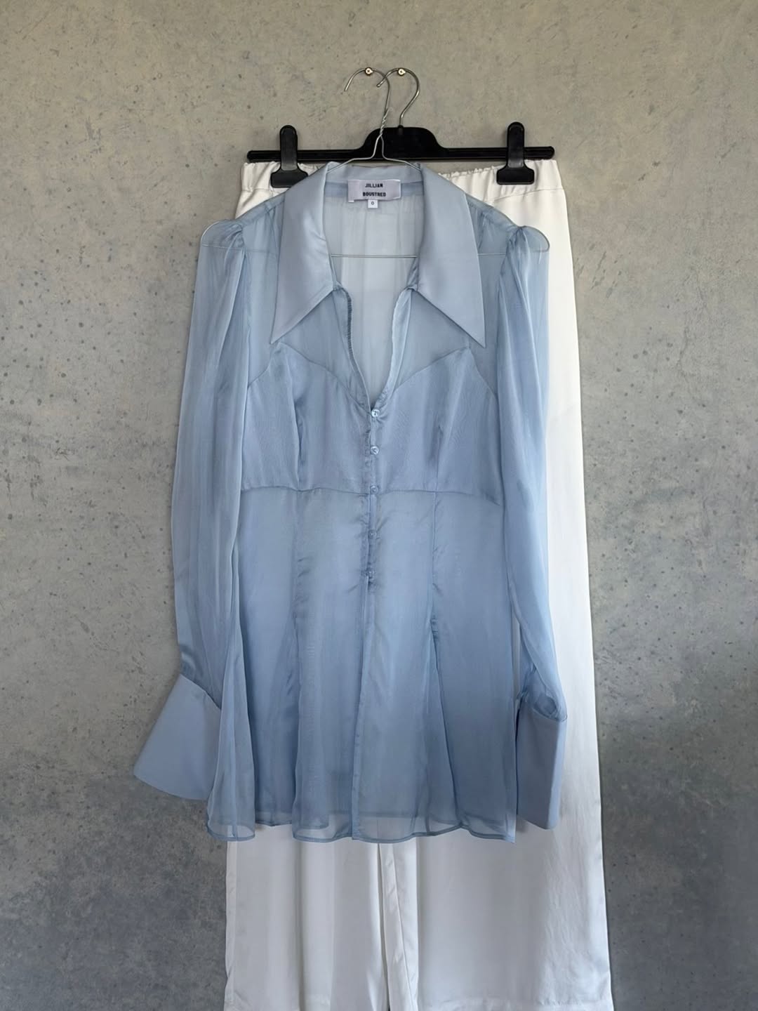a light blue long sleeve top hanging up with a long white skirt hanging behind it against a plain wall jillian boustred
