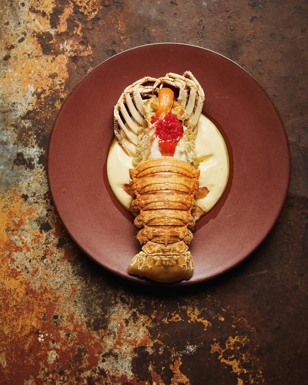 An elegantly plated salt and pepper bug from Ester, sitting atop a maroon ceramic dish and a marble table.