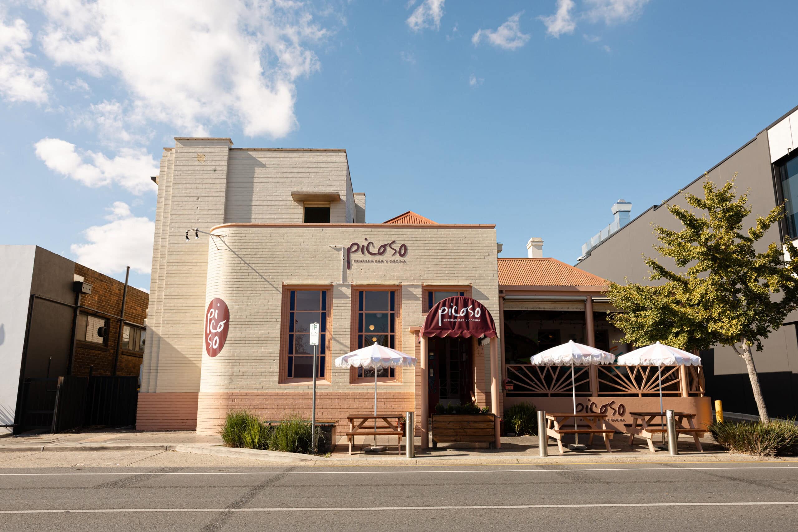 Street-facing exterior of Picoso Mexican Bar Y Cocina, Adelaide, a cream brick building with a burgundy awning, outdoor bench seating, and fringed umbrellas.
