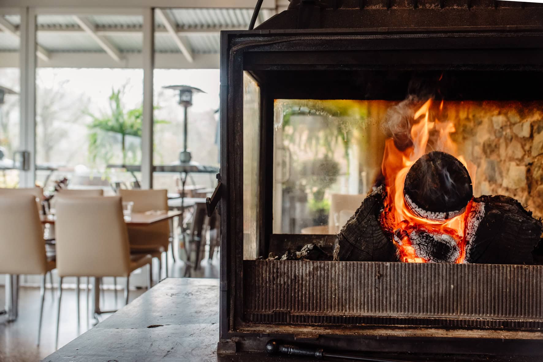 Close-up of a roaring wood fireplace at Inglewood Inn, Adelaide, with a light-filled dining room and garden views in the background.
