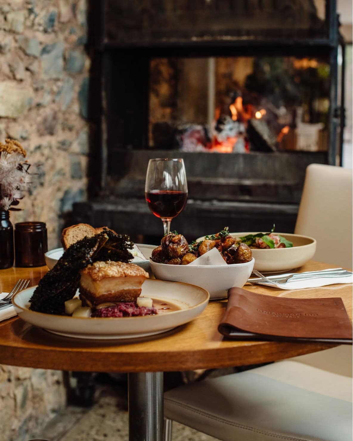 A spread of winter dishes and a glass of red wine on a timber table beside a crackling stone fireplace at Inglewood Inn, Adelaide.