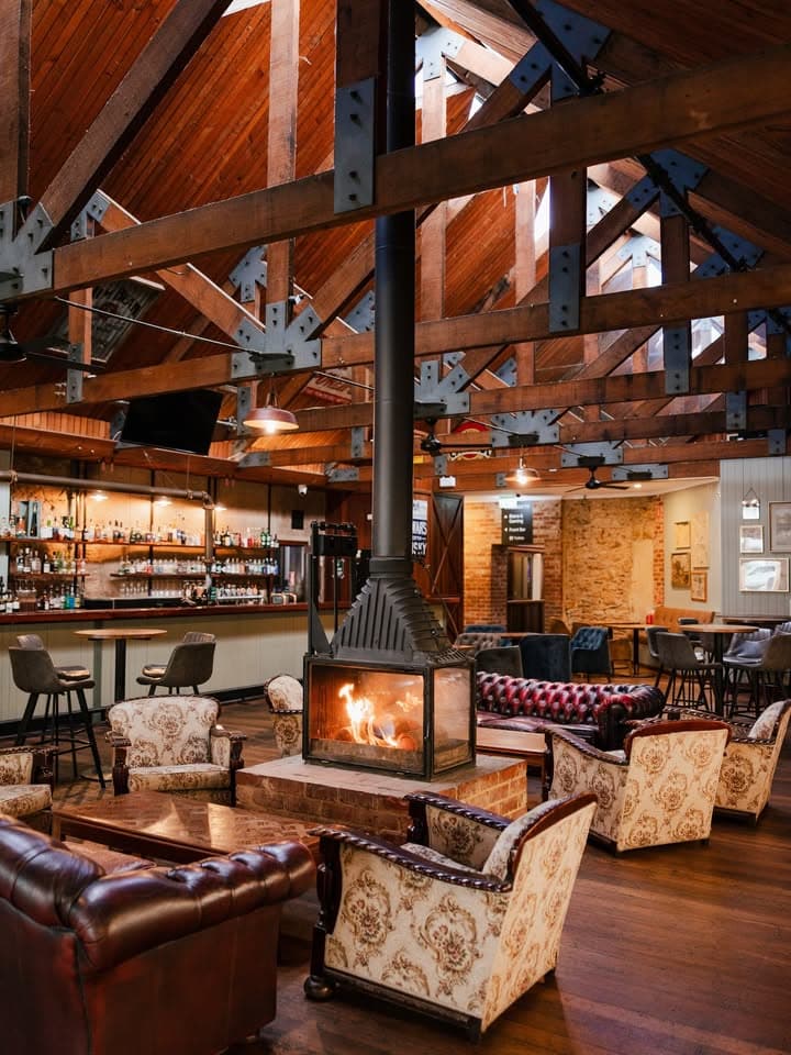 Spacious bar at The Gully Public House & Garden, Adelaide, featuring a central freestanding fireplace, leather Chesterfield sofas, floral armchairs, and a soaring timber ceiling with exposed steel trusses.