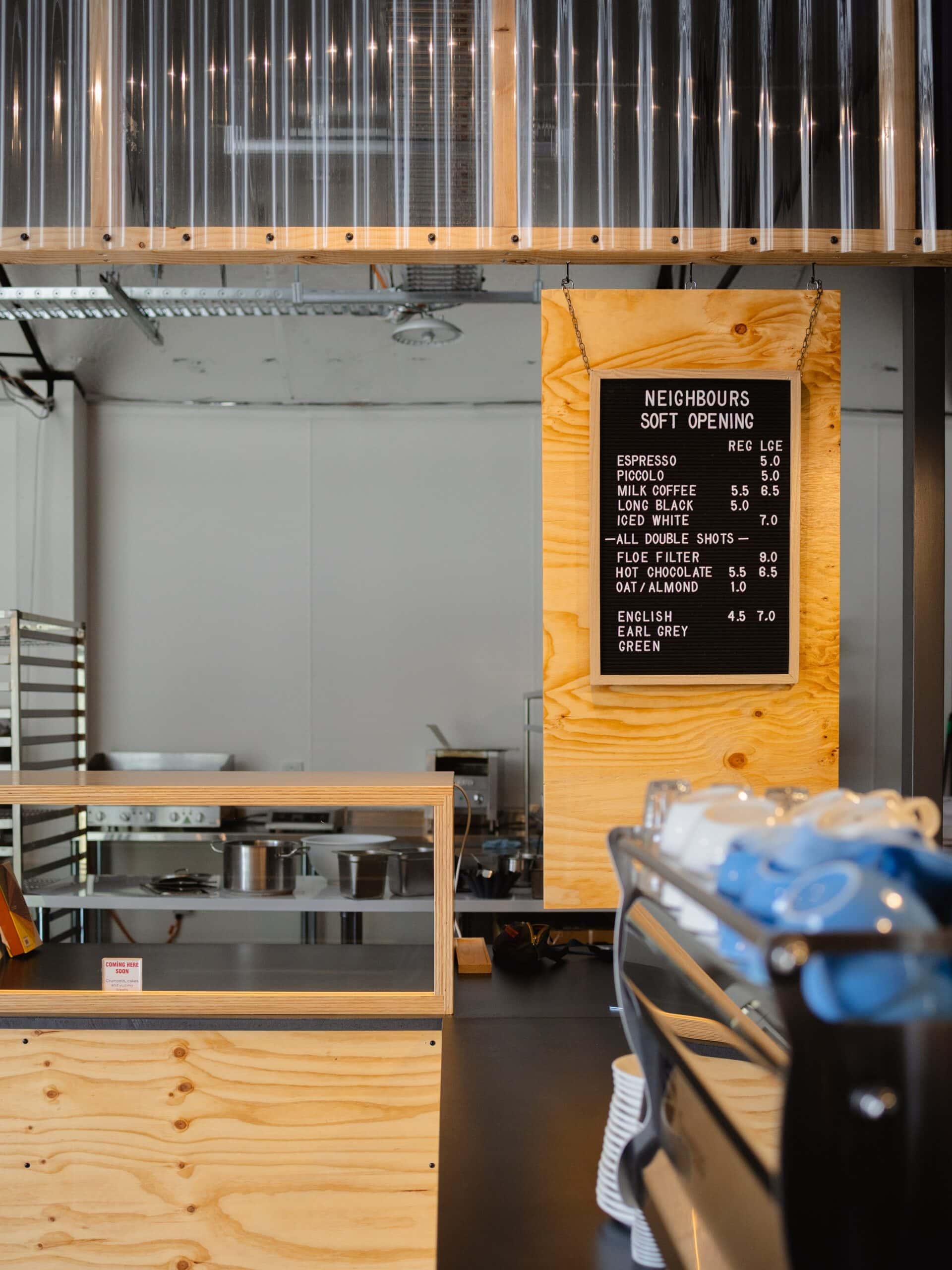 The counter interior of Neighbours café in McLaren Vale during soft opening, with a black letterboard drinks menu suspended from a raw plywood wall listing espresso, filter coffee, and teas, an espresso machine stacked with blue-rimmed cups, and a view through to the open kitchen.