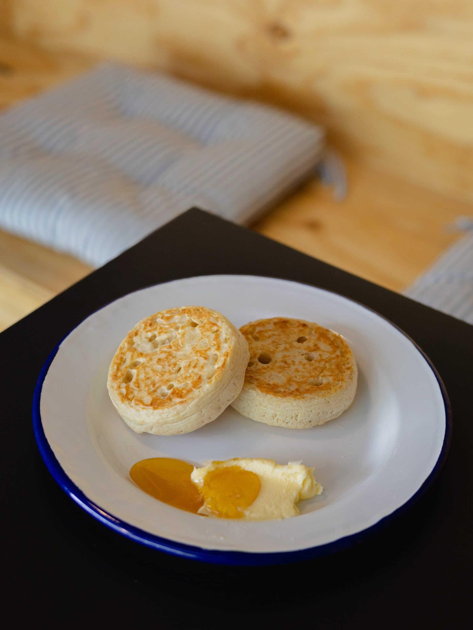 Two toasted sourdough crumpets from Neighbours in McLaren Vale served on a white enamel plate with a blue rim, accompanied by butter and honey, on a dark table with striped linen napkins and a pine wood surface in the background.