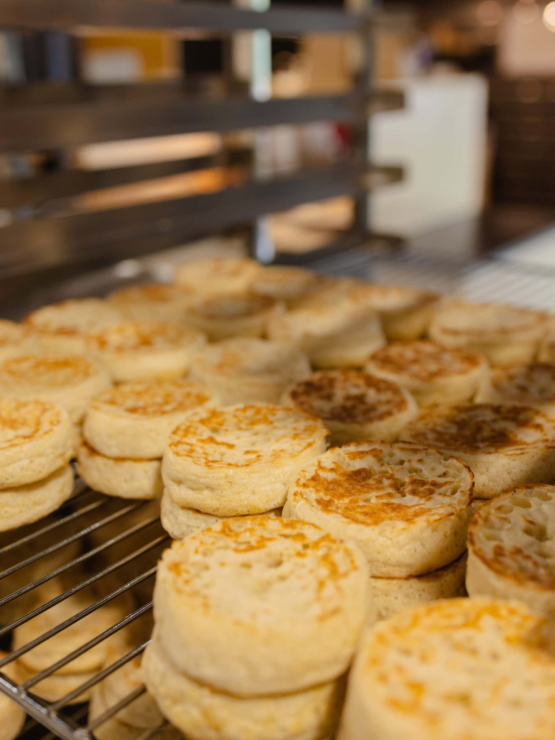 Dozens of freshly made sourdough crumpets cooling in stacks on a wire rack in the Neighbours kitchen, their golden-toasted tops showing the characteristic bubbled, holey texture of a well-fermented sourdough crumpet.