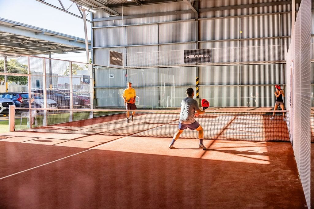 12:30 PM Three players competing on an indoor padel court at Padel Fit Adelaide, with HEAD-branded signage and a corrugated iron warehouse backdrop.