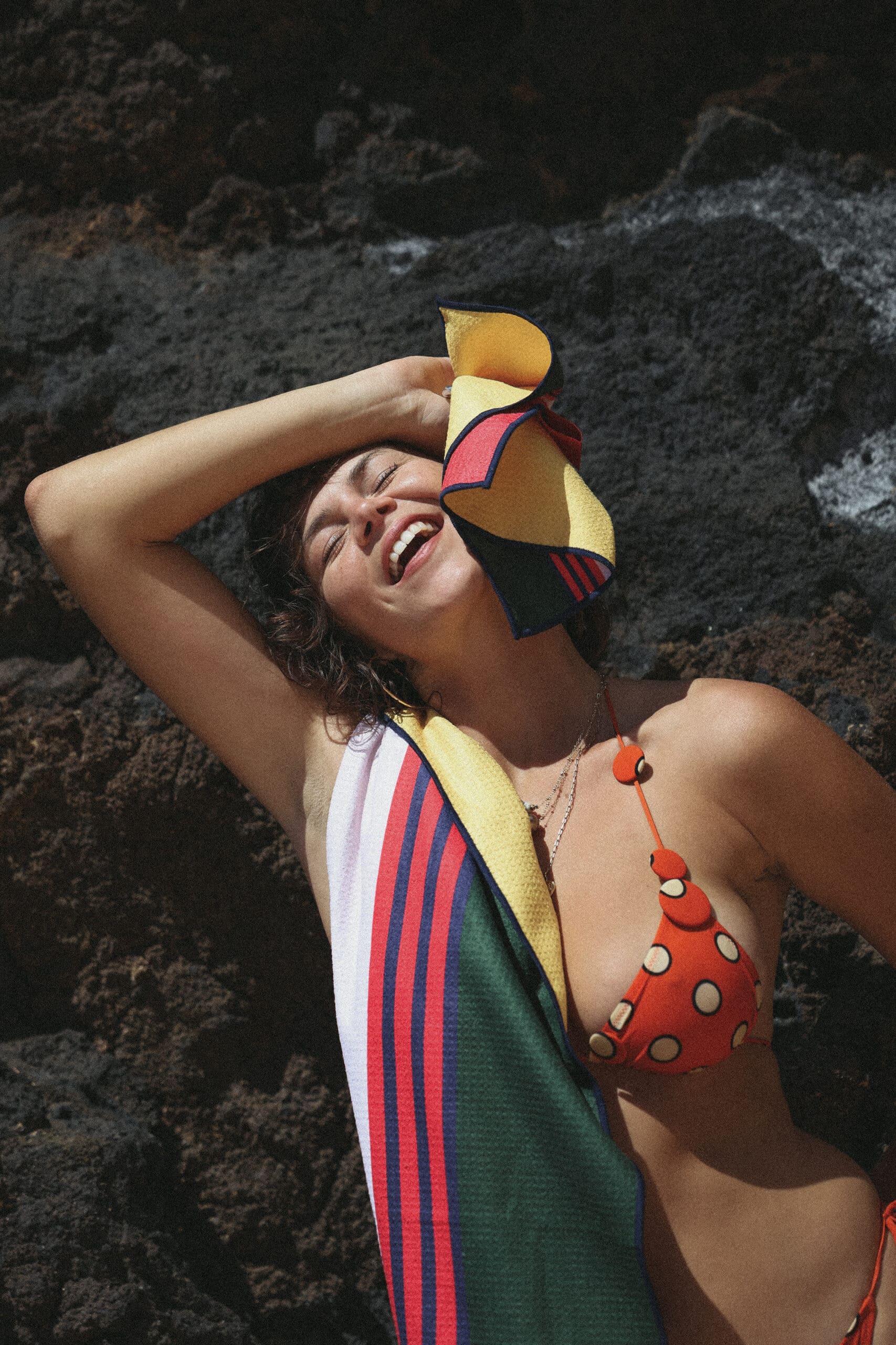 Model laughing with Clammy multicolour stripe waffle-weave towel draped over shoulder against coastal rocks