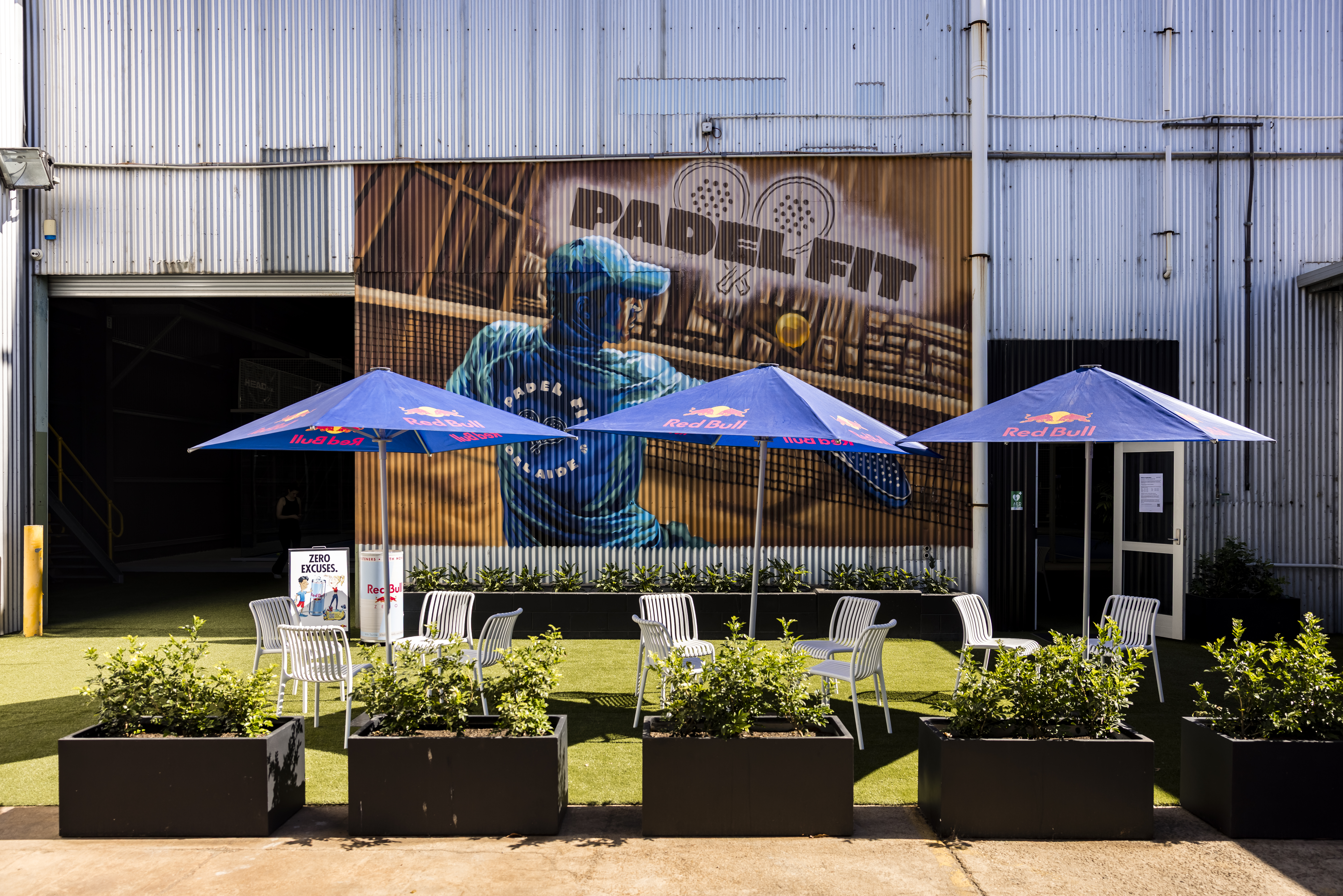 11:57 AM Outdoor seating area at Padel Fit Adelaide, with Red Bull umbrellas, white chairs, and planter boxes in front of a warehouse mural featuring a padel player.