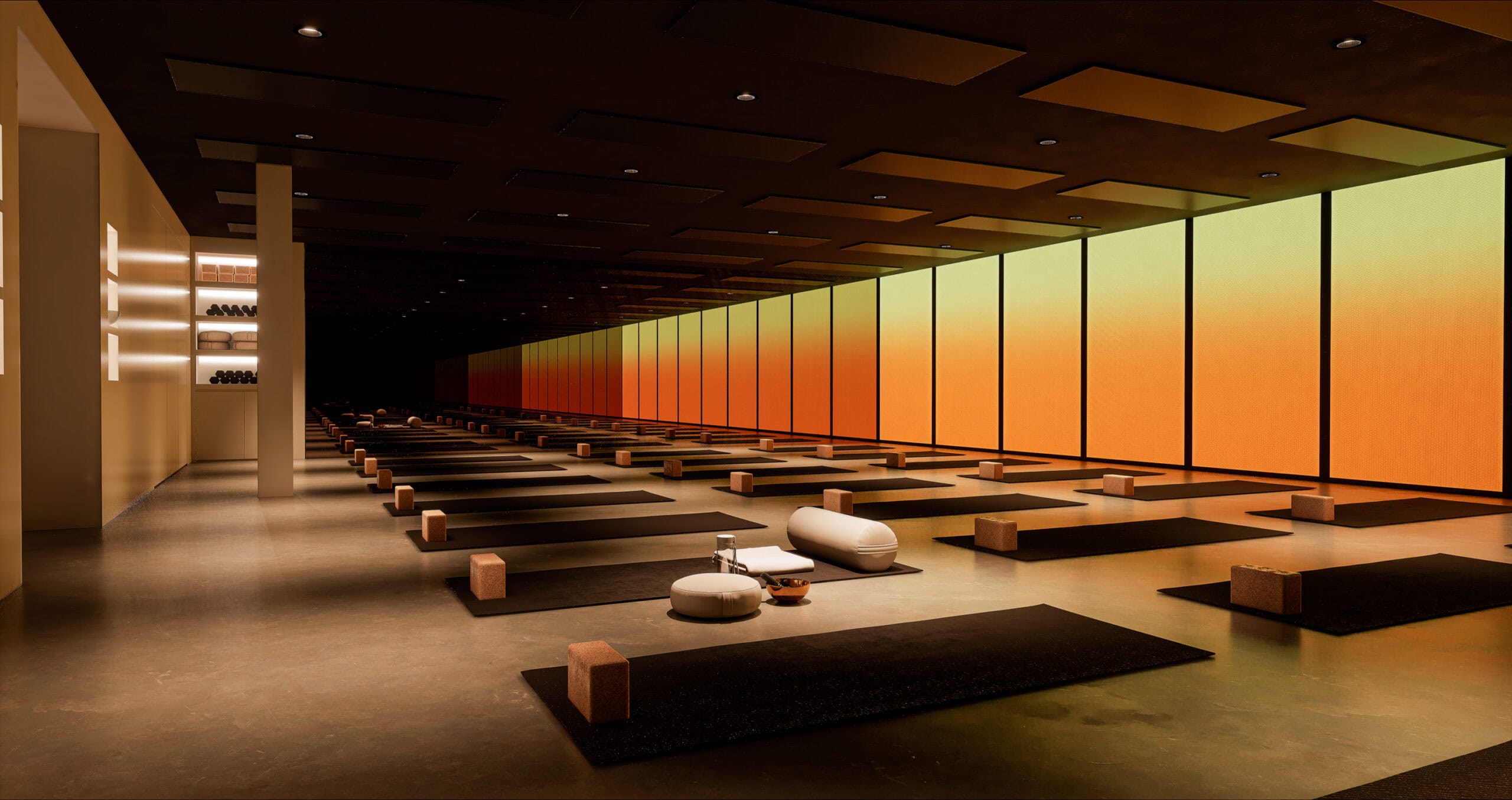 A render of Level Eleven's yoga studio with rows of dark mats and cork blocks set against floor-to-ceiling amber and burnt orange gradient-lit windows.