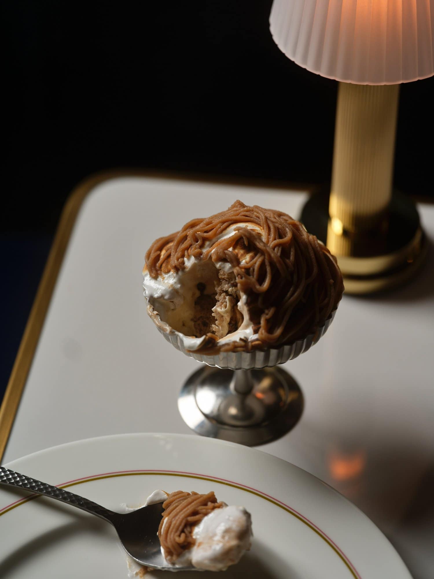 A Mont Blanc–style bombe alaska dessert served in a silver coupe glass, cut open to reveal layers of meringue, cream, and cake beneath chestnut vermicelli piped in curling threads. A spoonful of the dessert rests on a gold-rimmed white plate in the foreground, with a small glowing lamp behind on a marble table.