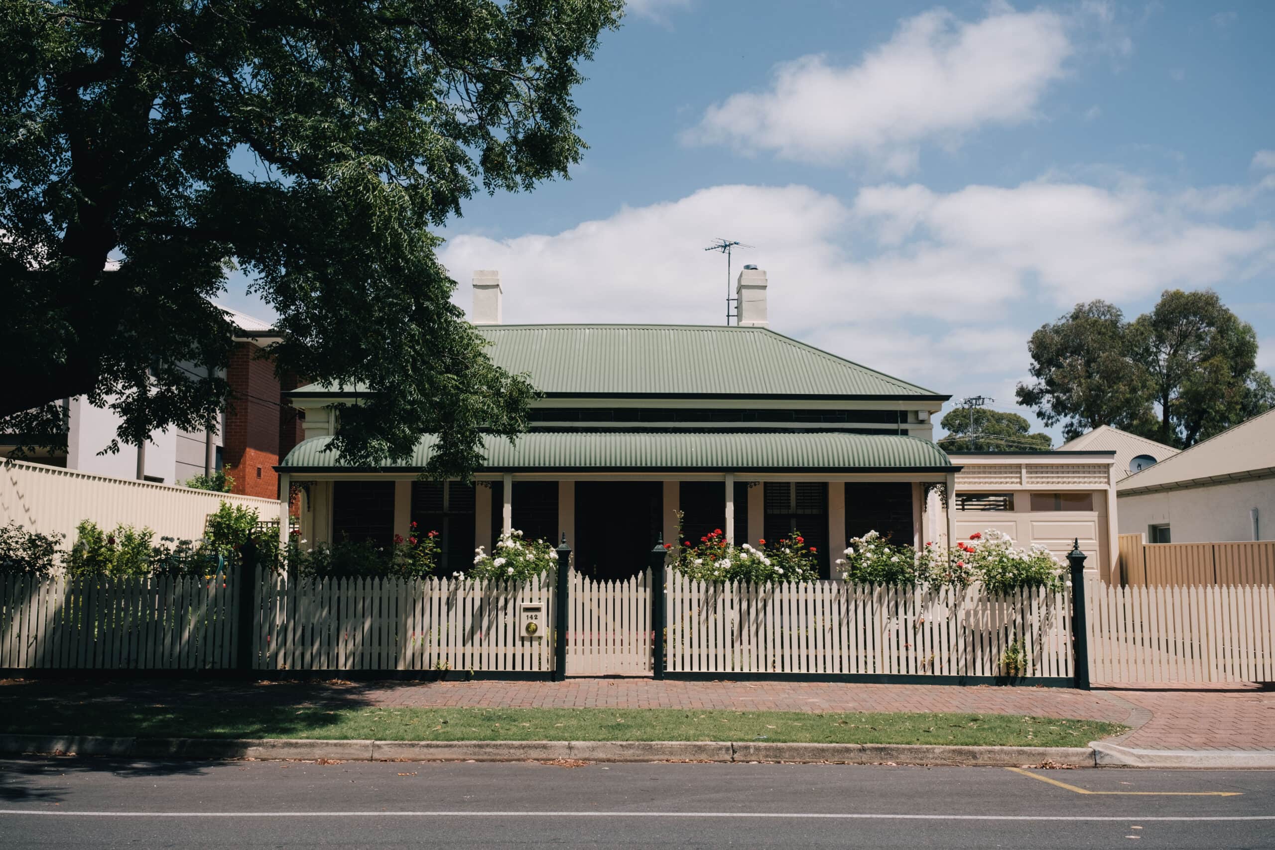A charming heritage cottage in Norwood, Adelaide featuring a green corrugated iron roof, white picket fence and rose garden in full bloom.