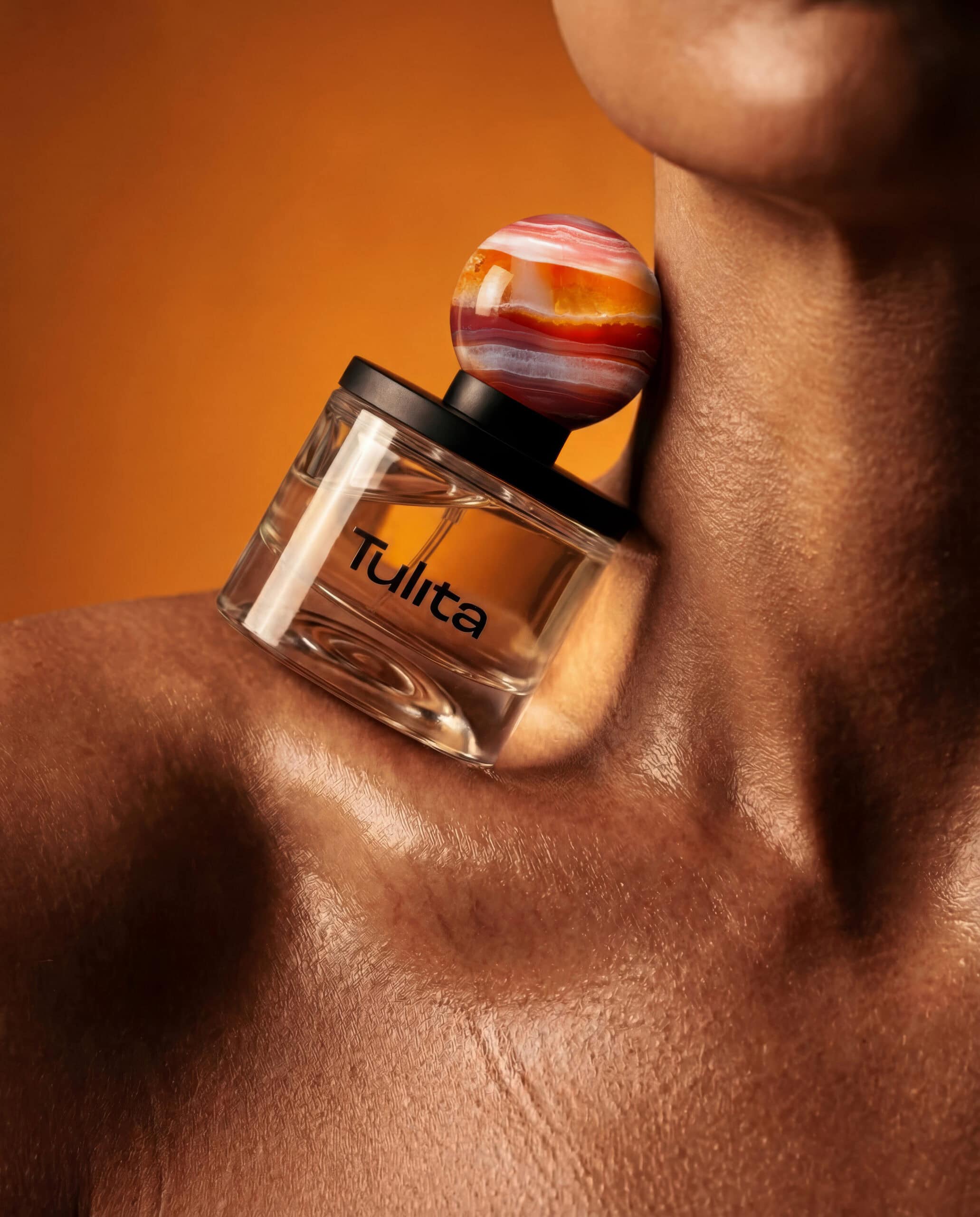Tulita Kama perfume bottle with carnelian marble stopper resting against a model's shoulder against an orange background.