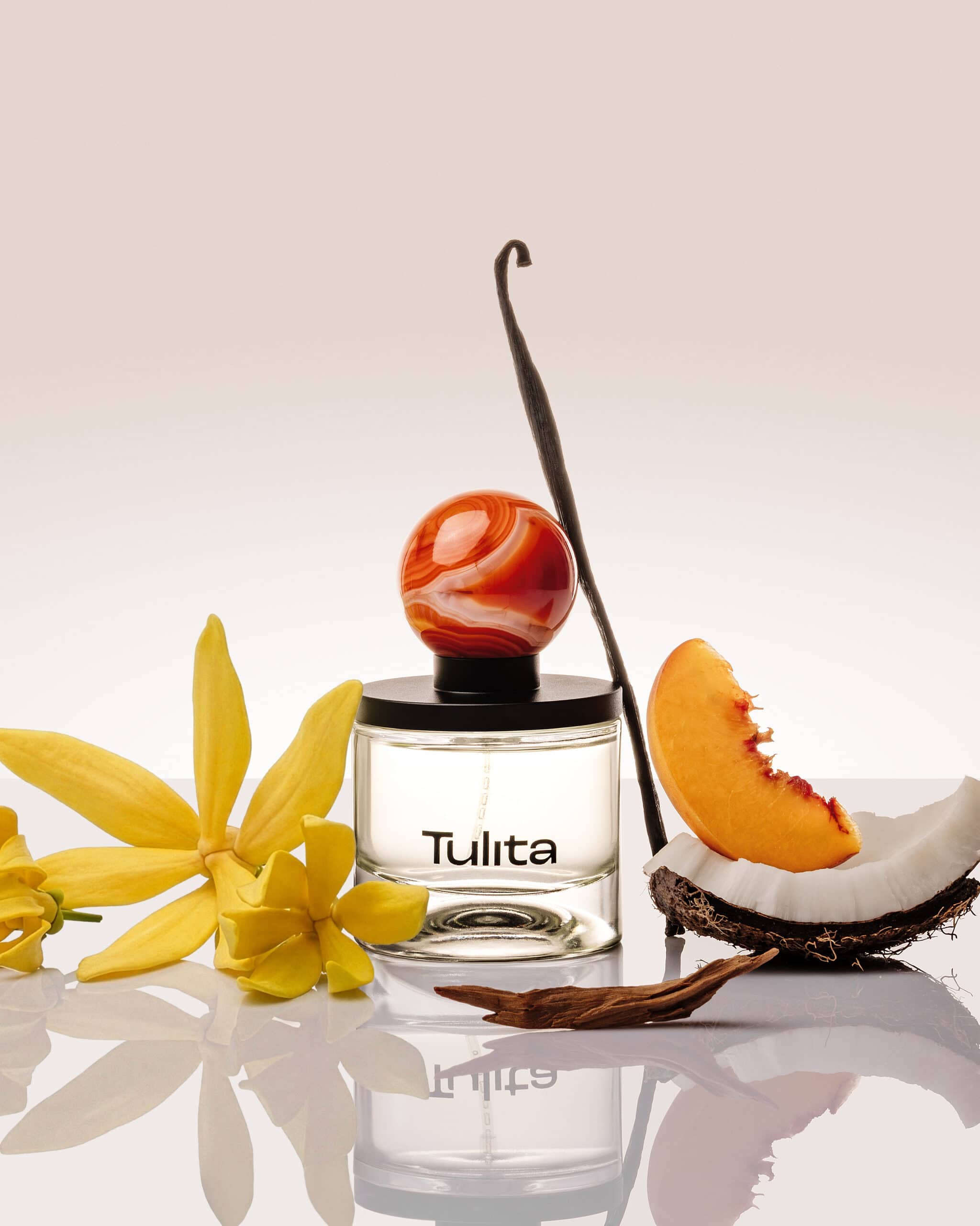 Tulita Kama perfume bottle with carnelian marble stopper surrounded by ylang ylang flowers, vanilla bean, peach slice and coconut on a reflective white surface