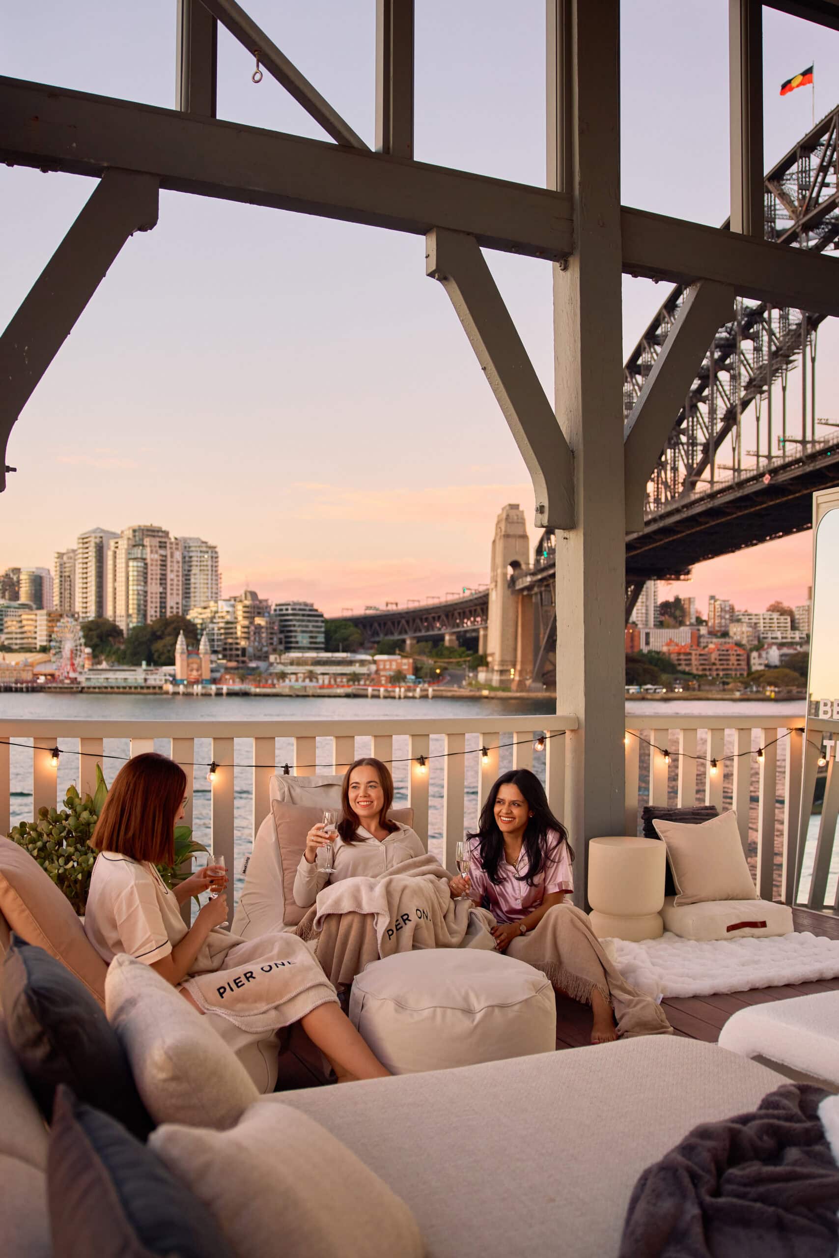 Three friends in pyjamas toasting champagne on Harbourside Cinema balcony with Sydney Harbour Bridge and Luna Park at sunset at Pier One Walsh Bay Sydney