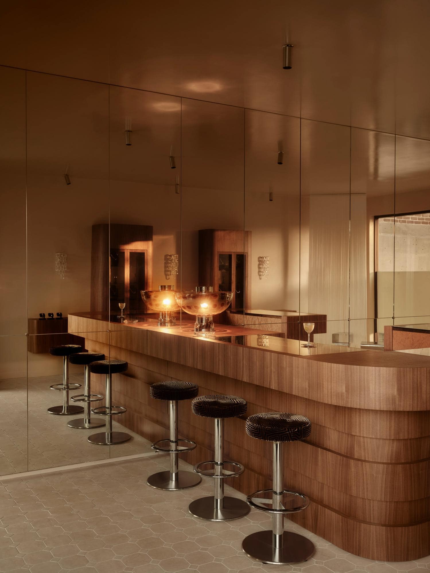 Jewellery store bar area with long curved walnut counter, row of bar stools with geometric patterned upholstery and chrome bases, bronze-toned mirror wall and ceiling creating a warm intimate atmosphere