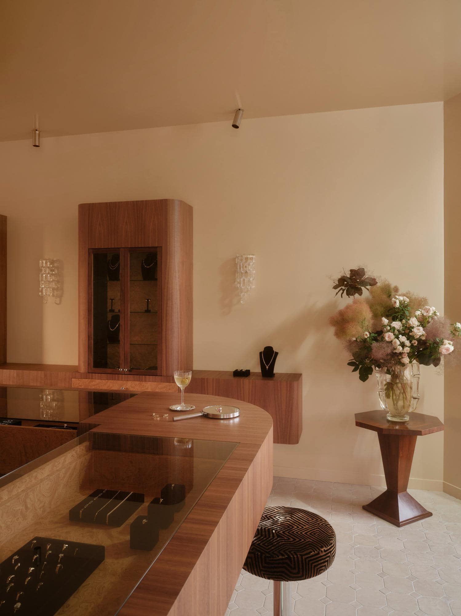 Jewellery store interior showing curved walnut service counter with glass of white wine and silver tray, wall-mounted display shelf, glass cabinet with necklaces, and large floral arrangement on a pedestal side table