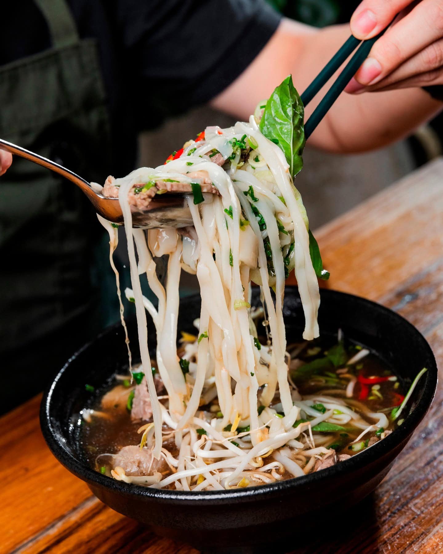 Someone using a spoon and chopsticks to pick up the rice noodles in a bowl of pho at Hello Auntie.