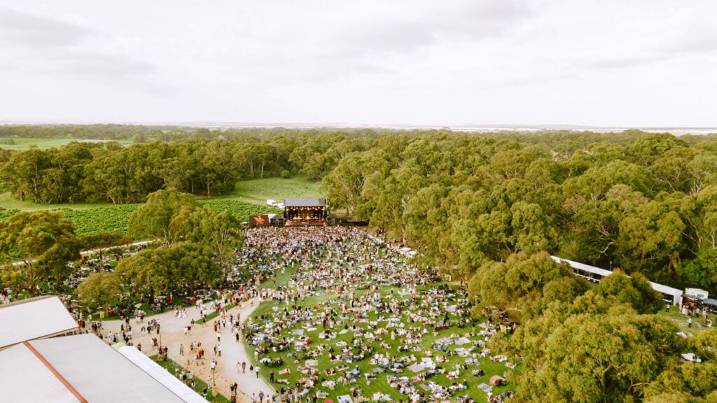 Aerial view of Handpicked Festival at Lake Breeze Wines, with hundreds of festival-goers gathered on picnic rugs beneath towering gum trees and a live music stage in the distance.