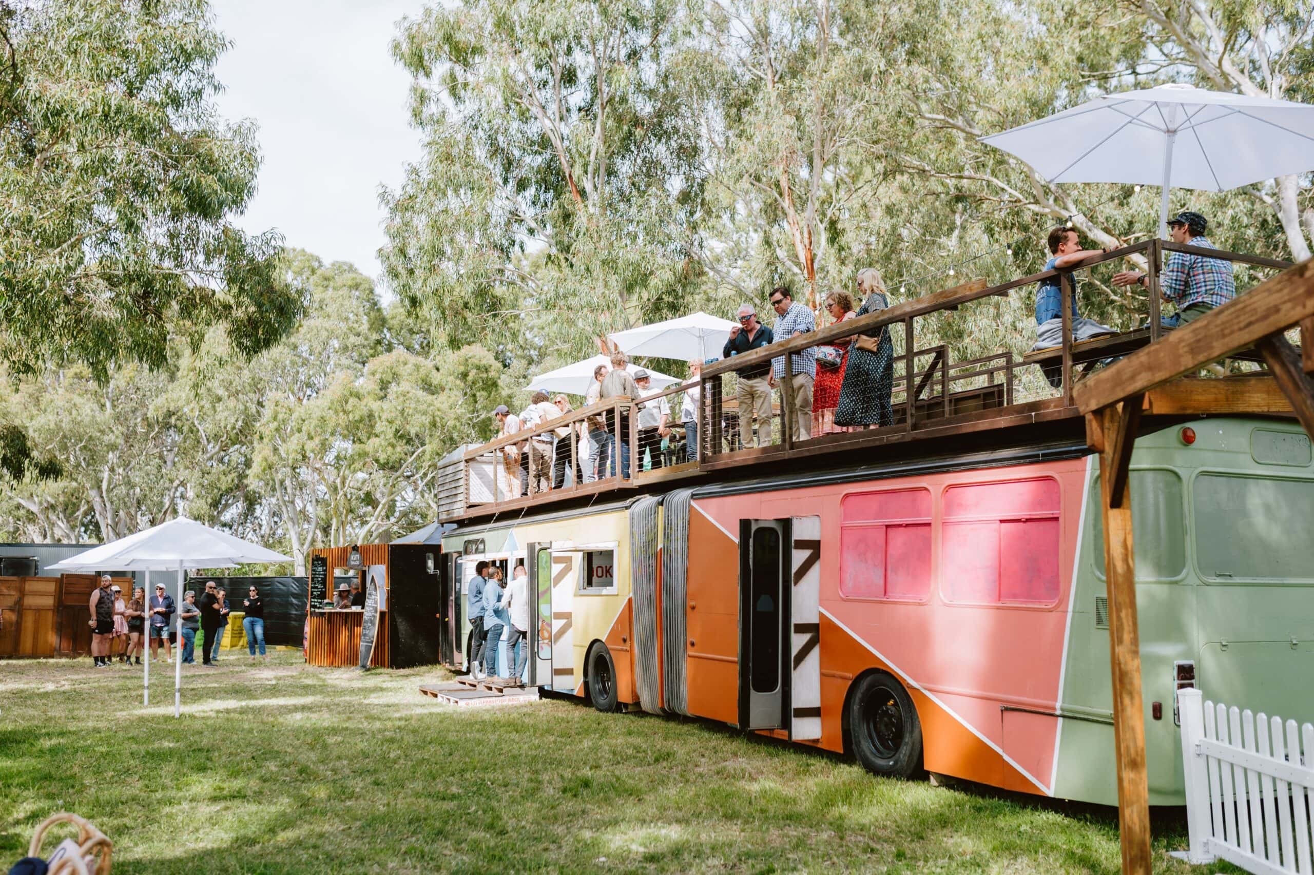 Festival guests relaxing on an elevated viewing deck built atop a colourful converted bus, surrounded by gum trees at Handpicked Festival.