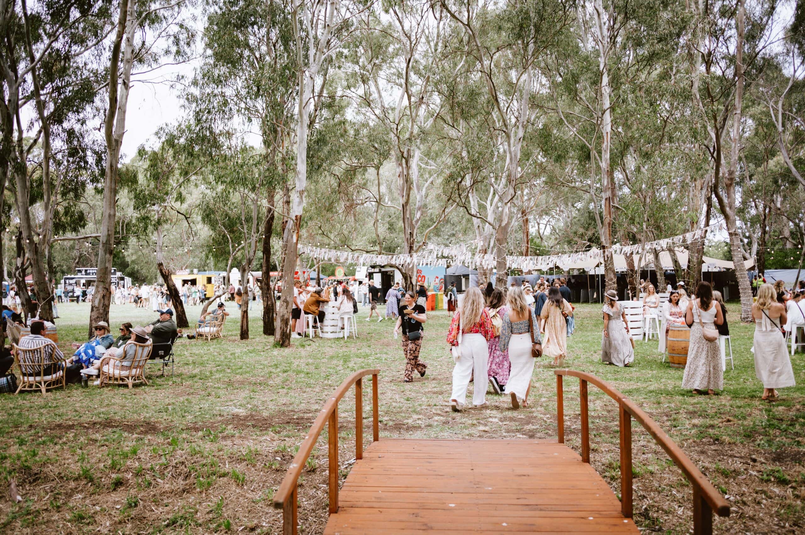 Festival-goers wandering through a shaded woodland precinct lined with food vendors, bars and seating areas at Handpicked Festival.