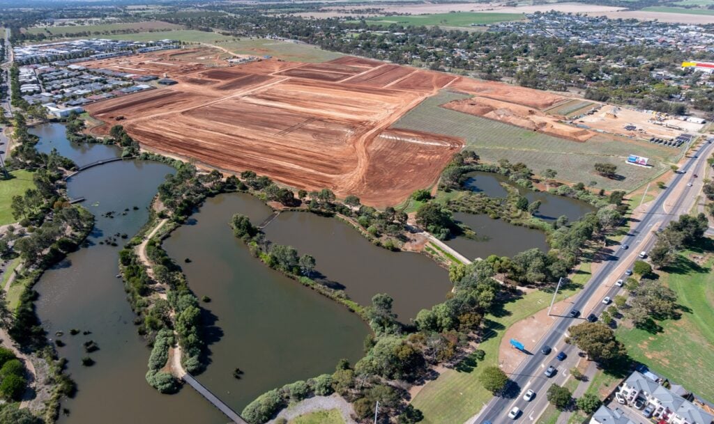 Aerial view of the Playford Alive first homebuyers precinct in Adelaide's northern suburbs, showing civil works underway on a large parcel of red earth land, surrounded by wetlands, parklands and established residential streets.
