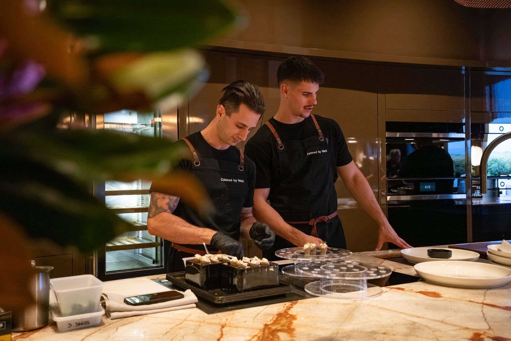 Two Catered by Matt chefs carefully plating canape portions at a marble kitchen bench during an evening event
