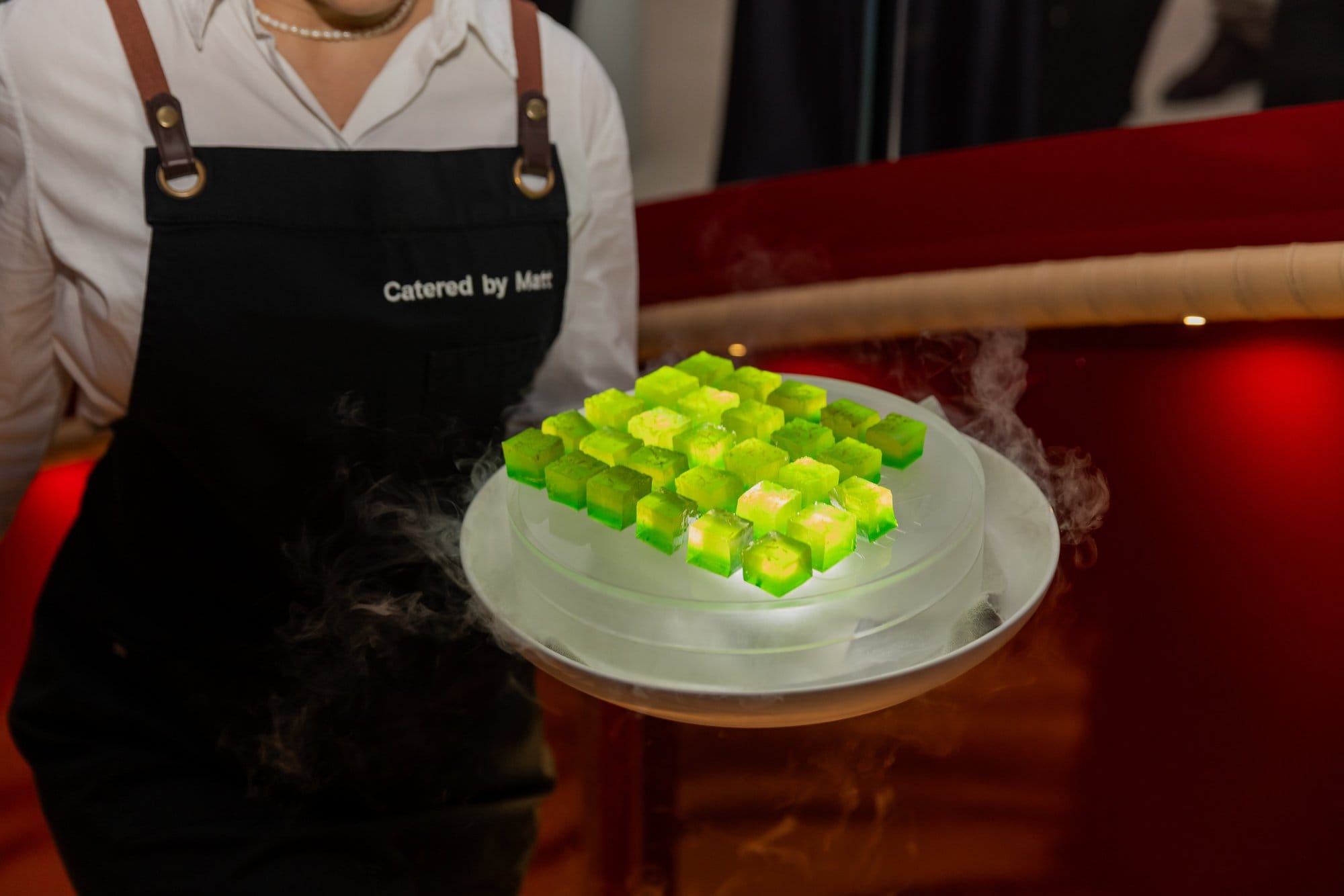 A Catered by Matt waiter presenting a dramatic platter of bright green jelly cubes with dry ice smoke at a formal event