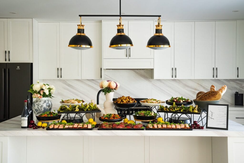 A full Catered by Matt buffet display across a marble kitchen island, with tiered black serving dishes, fresh flowers, wine and a printed menu card