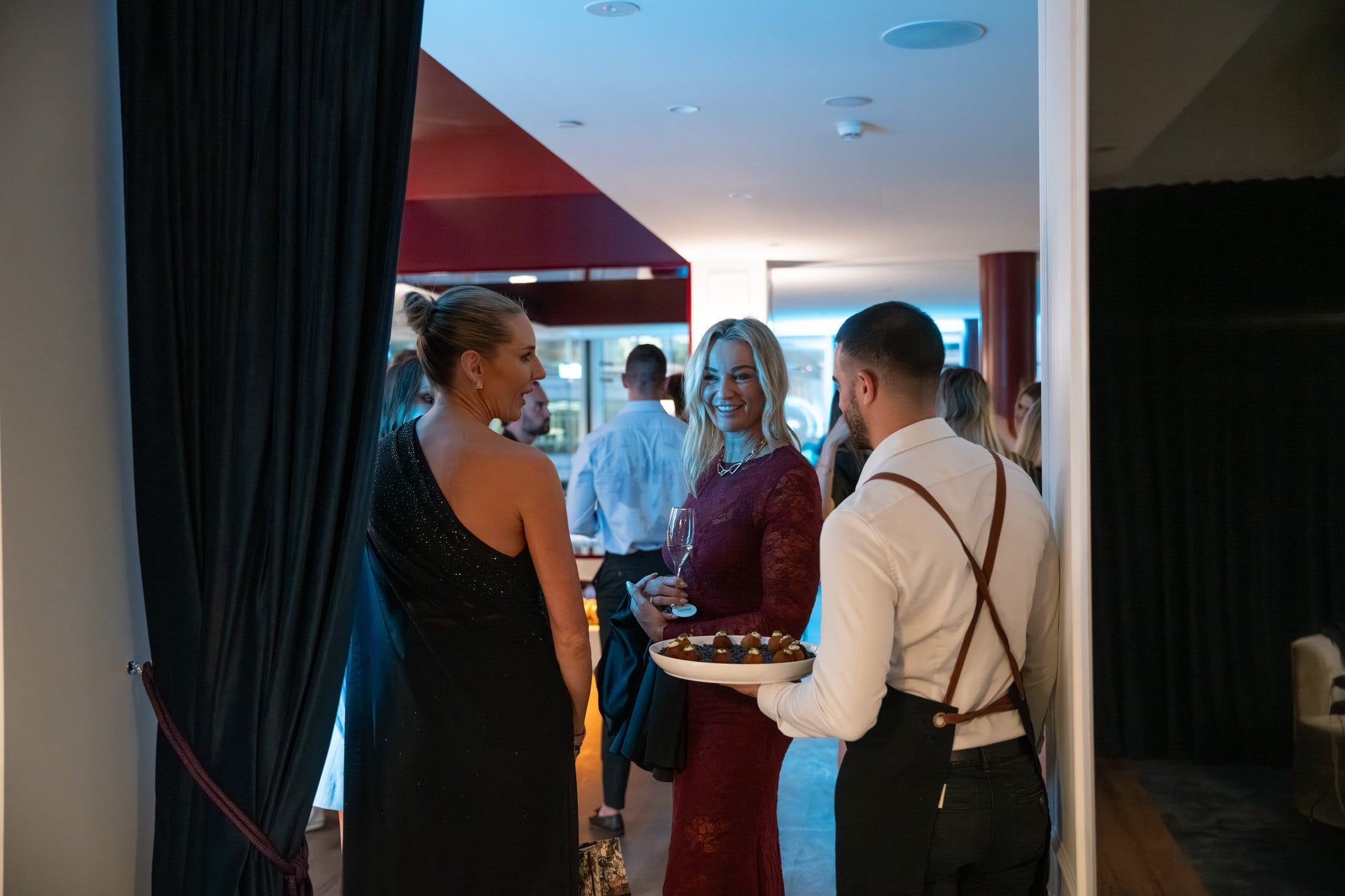 A Catered by Matt waiter in a white shirt and leather apron offering roaming canapés to guests at a formal evening event