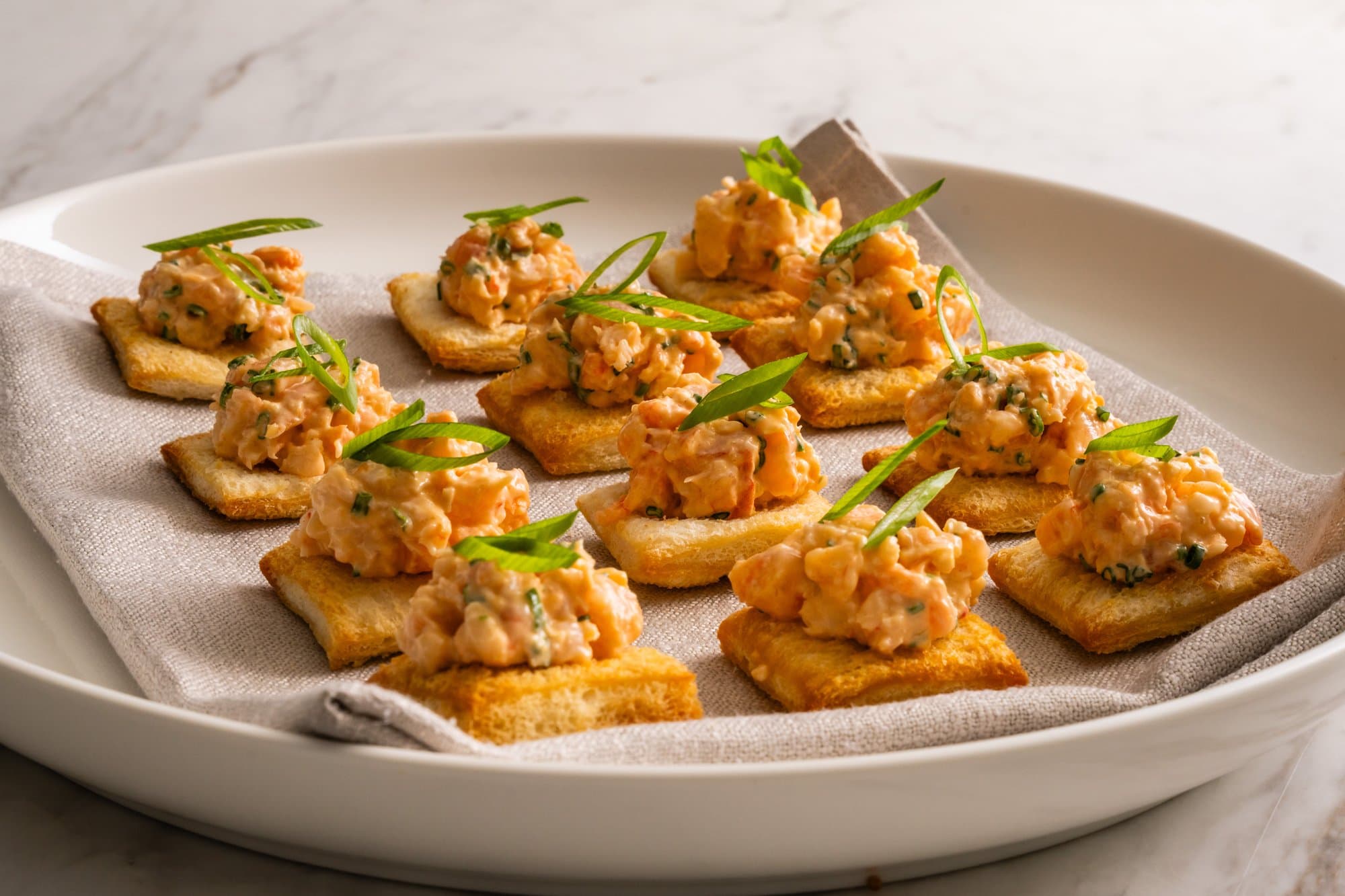 Prawn toast canapés topped with a creamy prawn mixture and fresh chives, arranged on a white plate with a linen napkin