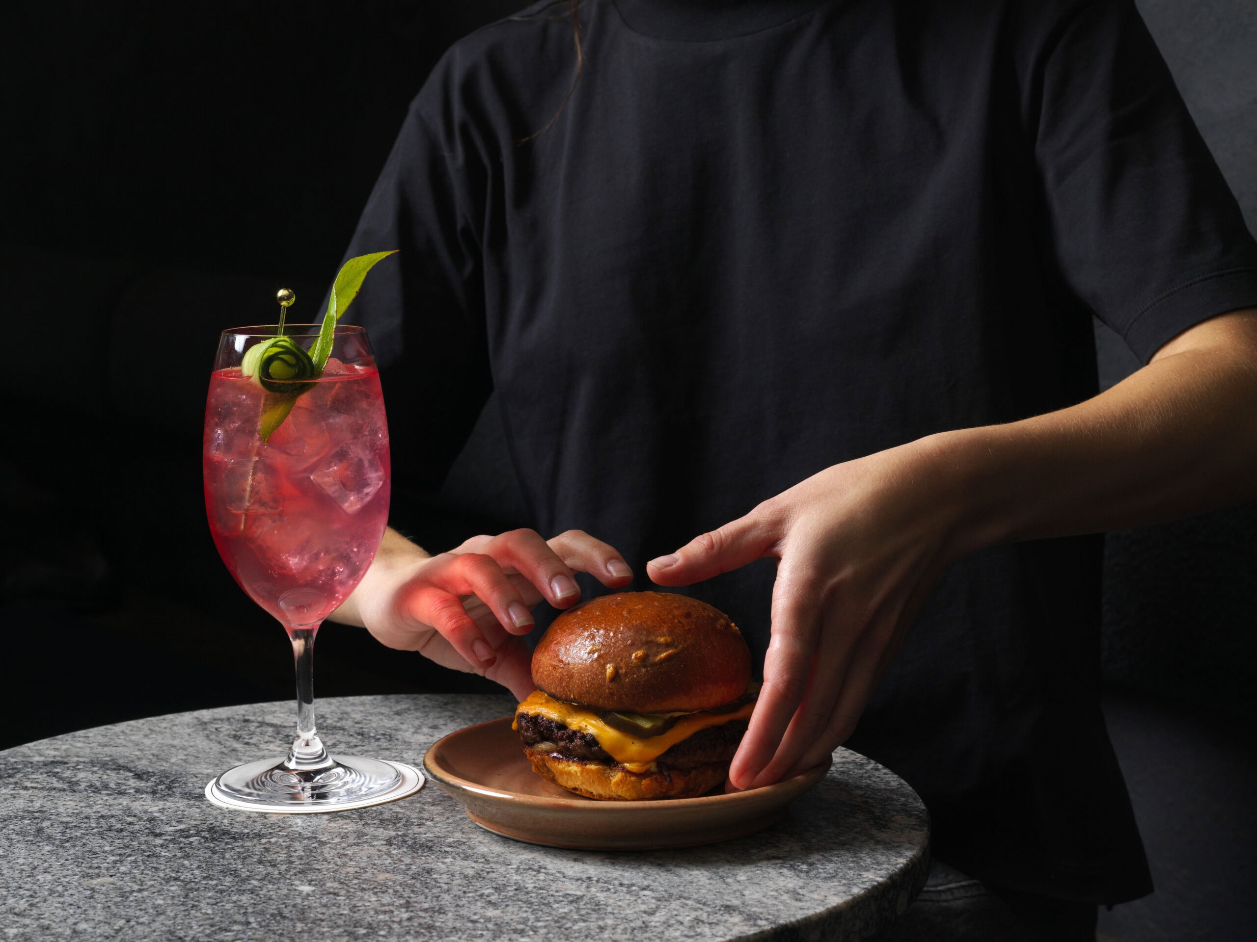 A server's hands presenting a cheeseburger on a ceramic plate alongside a crimson spritz cocktail garnished with a cucumber curl and fresh herbs in a wine glass, on a round granite table against a black background at arkhe in Adelaide.