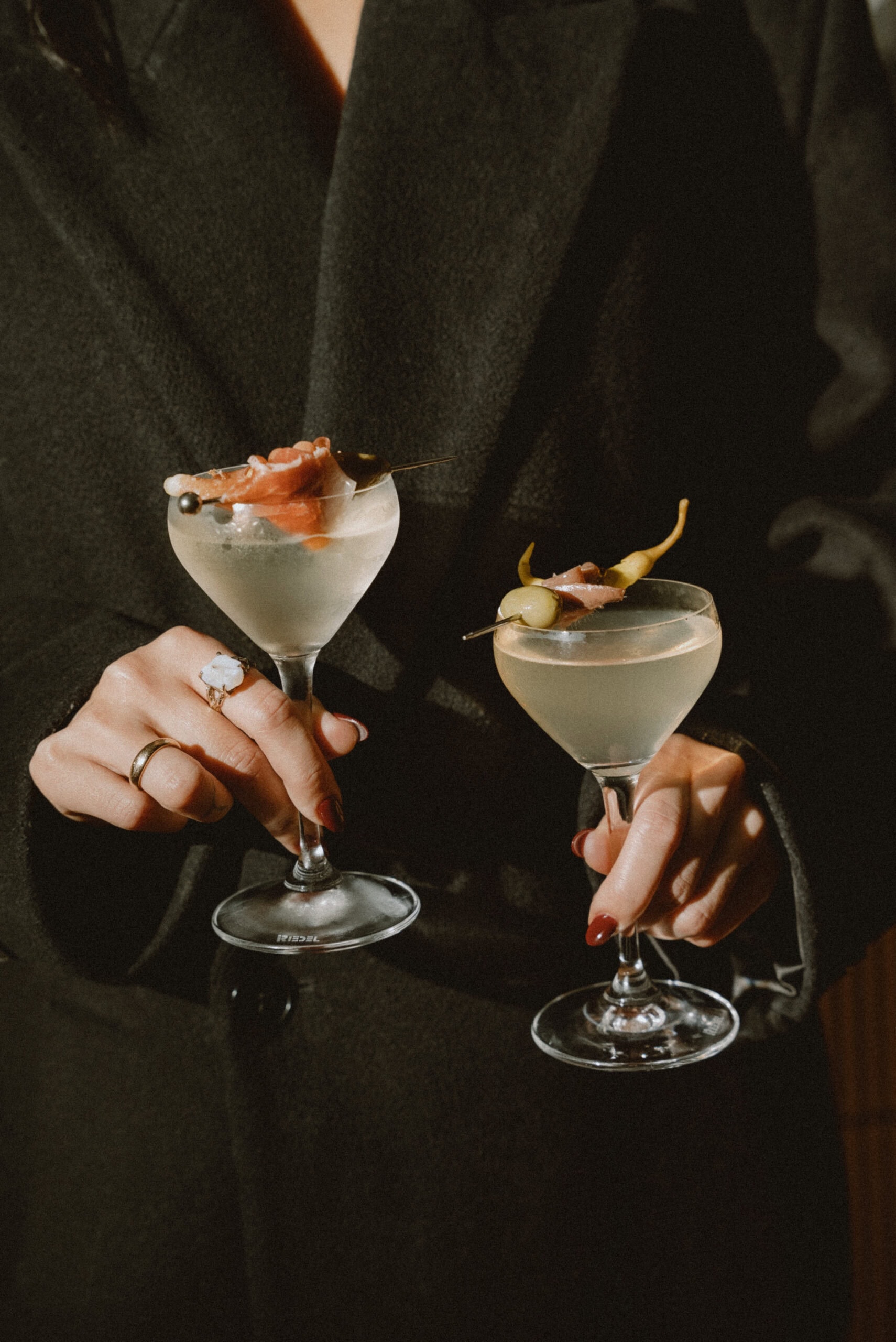 A moody, close-up shot of two people holding up cocktails in coupe glasses. Each drink is elegantly garnished with savory skewers featuring olives, peppers, and prosciutto.