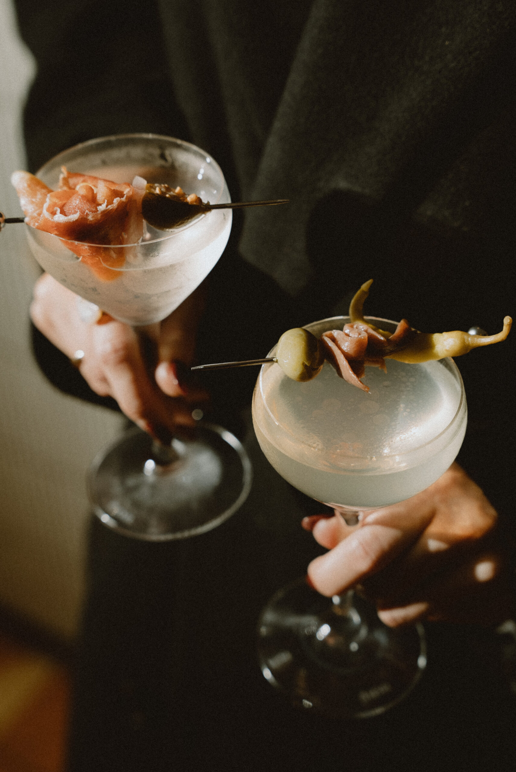 A moody, close-up shot of two people holding up cocktails in coupe glasses. Each drink is elegantly garnished with savory skewers featuring olives, peppers, and prosciutto.