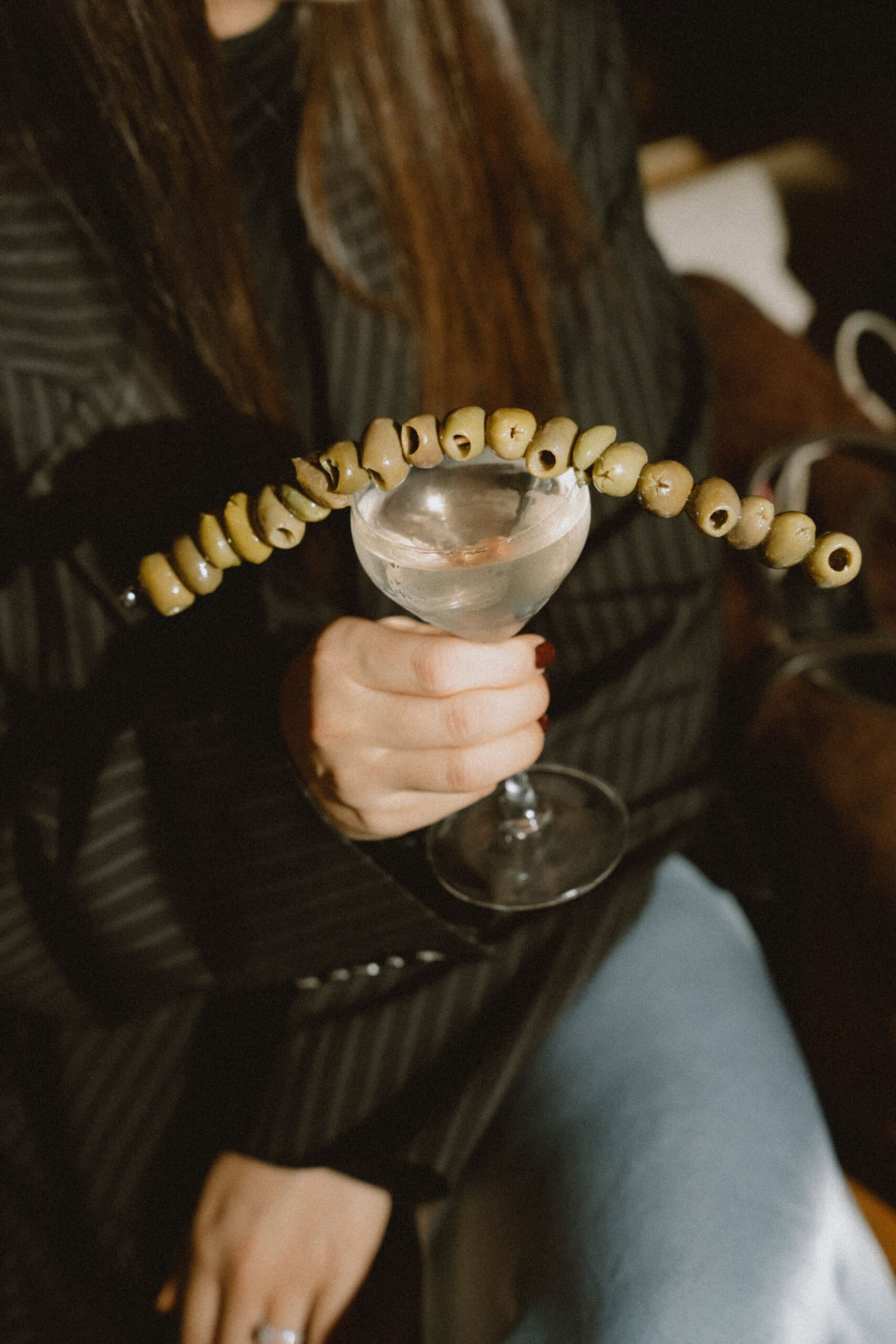 A close-up shot of a person wearing a pinstripe blazer holding a coupe cocktail glass. Balancing across the rim of the glass is an exceptionally long cocktail skewer stacked entirely with green olives, forming a wide arch.