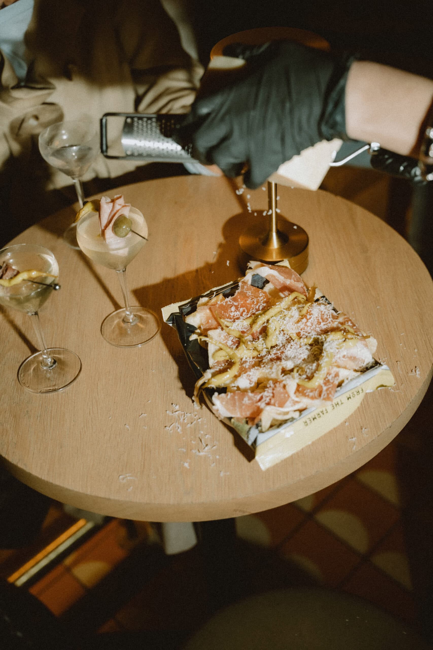 A top-down view of a round wooden bar table with three martini-style cocktails and a shared plate of prosciutto-topped flatbread. In the upper right, a hand in a black latex glove is actively grating fresh cheese over the food.