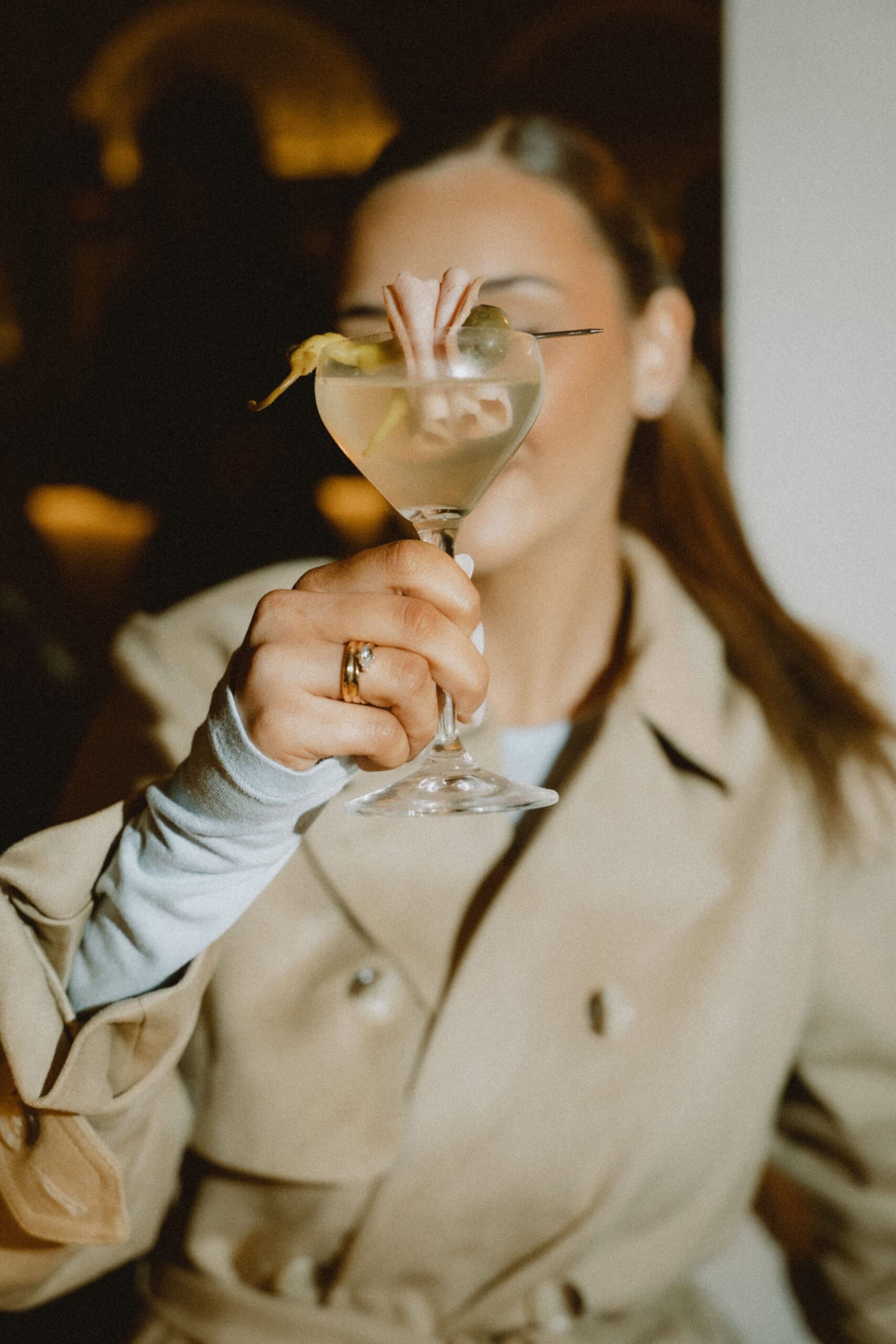 A person wearing a tan trench coat holds up a martini glass to partially obscure their face. The cocktail is garnished with an olive, a guindilla pepper, and a folded piece of charcuterie on a metal skewer.