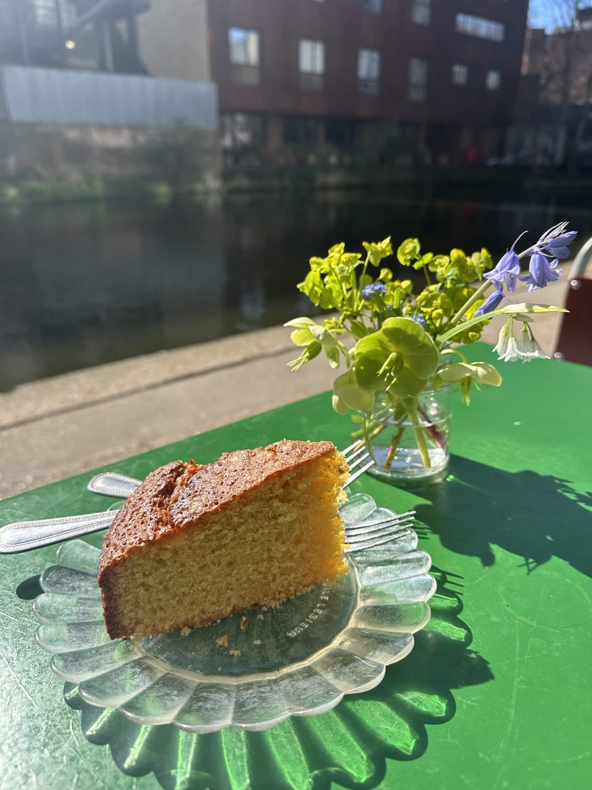 A homemade piece of cake on a glass plate with the canal in the background at tow path cafe in London.