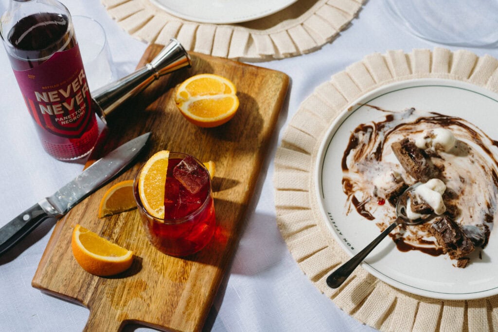 Never Never Panettone Negroni served over ice with an orange garnish, alongside the bottle and a dessert, styled on a wooden board.