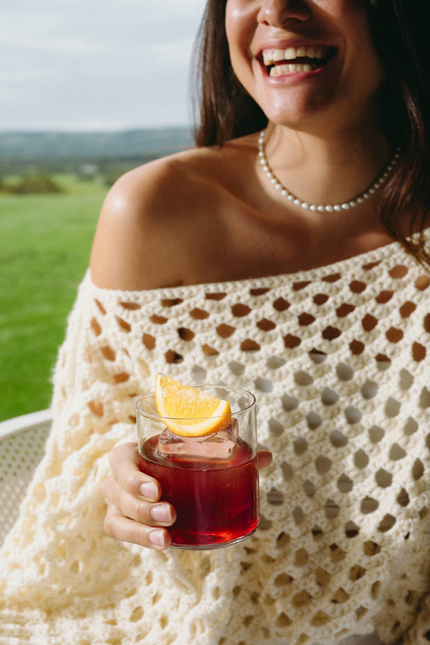 A woman smiles holding a Never Never Panettone Negroni over ice with an orange garnish, with green McLaren Vale vineyard views in the background.
