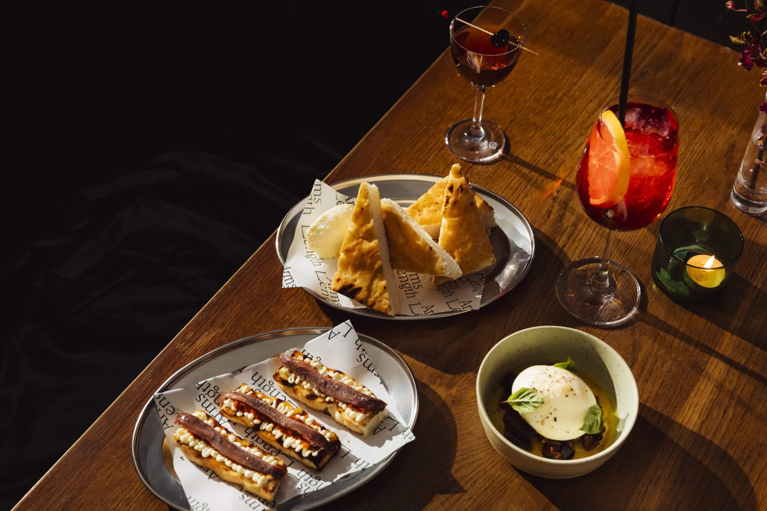 Anchovy toasts, burrata with basil, grilled focaccia with butter, a spritz and a Manhattan on a sunlit timber table at Arms Length bar, Potts Point Sydney