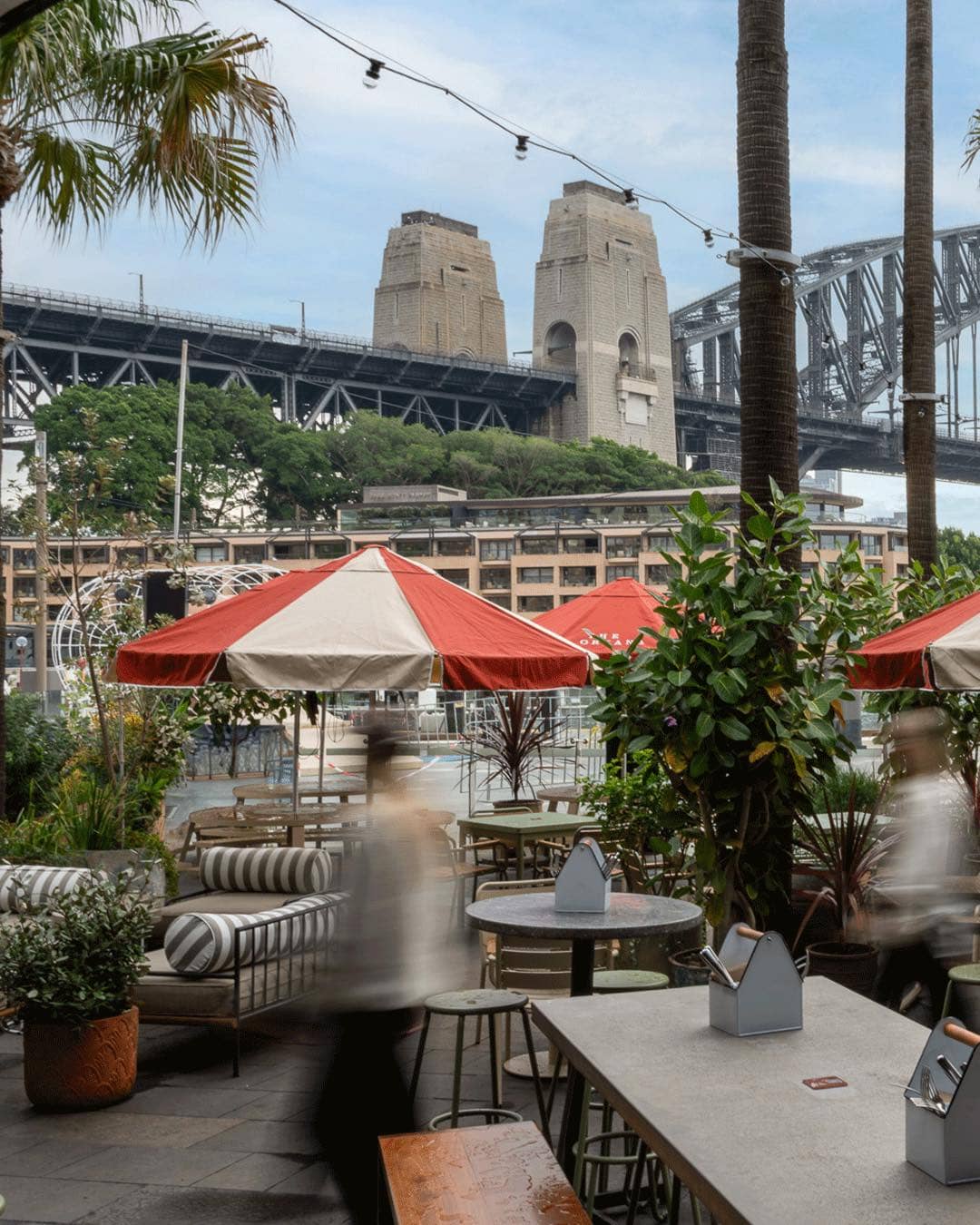 The beer garden at The Oriana, featuring orange and white striped umbrellas and a close-up view of the Sydney Harbour Bridge.