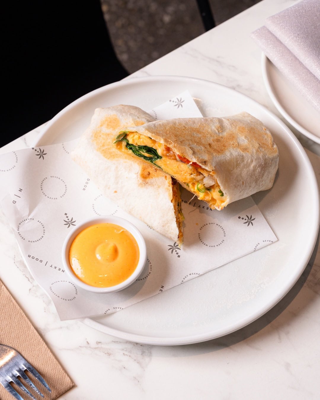 A scrambled egg breakfast wrap on a plate with a dish of sauce at next door in cronulla.
